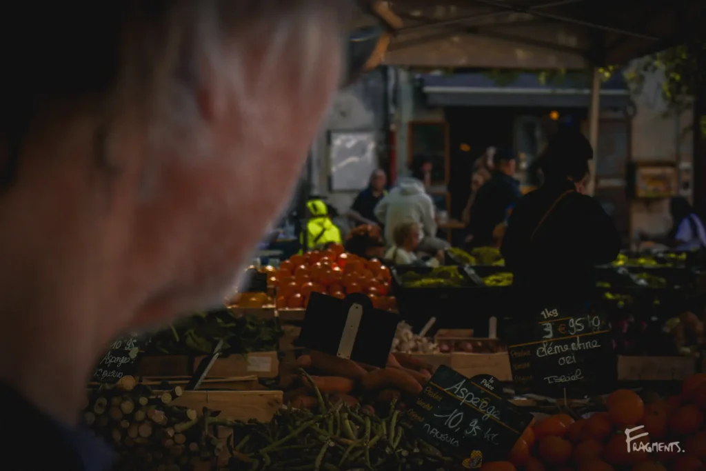 Passant au premier plan, au milieu des étals de fruits et légumes dans un marché à Montpellier